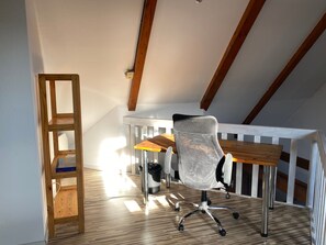 Une pièce avec un plafond en pente, un bureau en bois, une chaise de bureau à dossier en treillis métallique, une rambarde blanche et une échelle en bois.