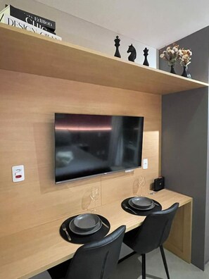 A modern dining area with a flat-screen TV mounted on a wooden wall, two black chairs, and a small table with plates and a wine glass. - Apartamento em Moema | Varanda e próximo do metrô (São Paulo)