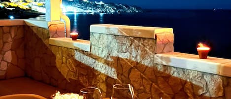 A table set for two with wine glasses, a candle, and a lantern, overlooking a coastal view at night.