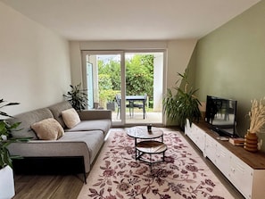 A living room with a sofa, a coffee table, a TV, and a sliding glass door leading to an outdoor area with a table and chairs. - Apartment with terrace and garden in Strasbourg (Strasbourg)