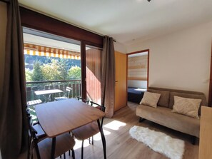 Una habitación con piso de madera, un sofá, una mesa con sillas, un balcón con vista a árboles y montañas, y una puerta que da a otra habitación.