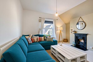 A cozy living room with a teal sofa, a white coffee table, a fireplace, and a window with curtains. - Host & Stay | Indigo Snug (Cromer)