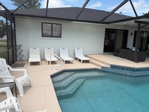 A swimming pool with a tiled edge, surrounded by white lounge chairs, and a covered area with a window. - Amazing Canal Home, Heated Pool, Spa & Fishing (Cape Coral)