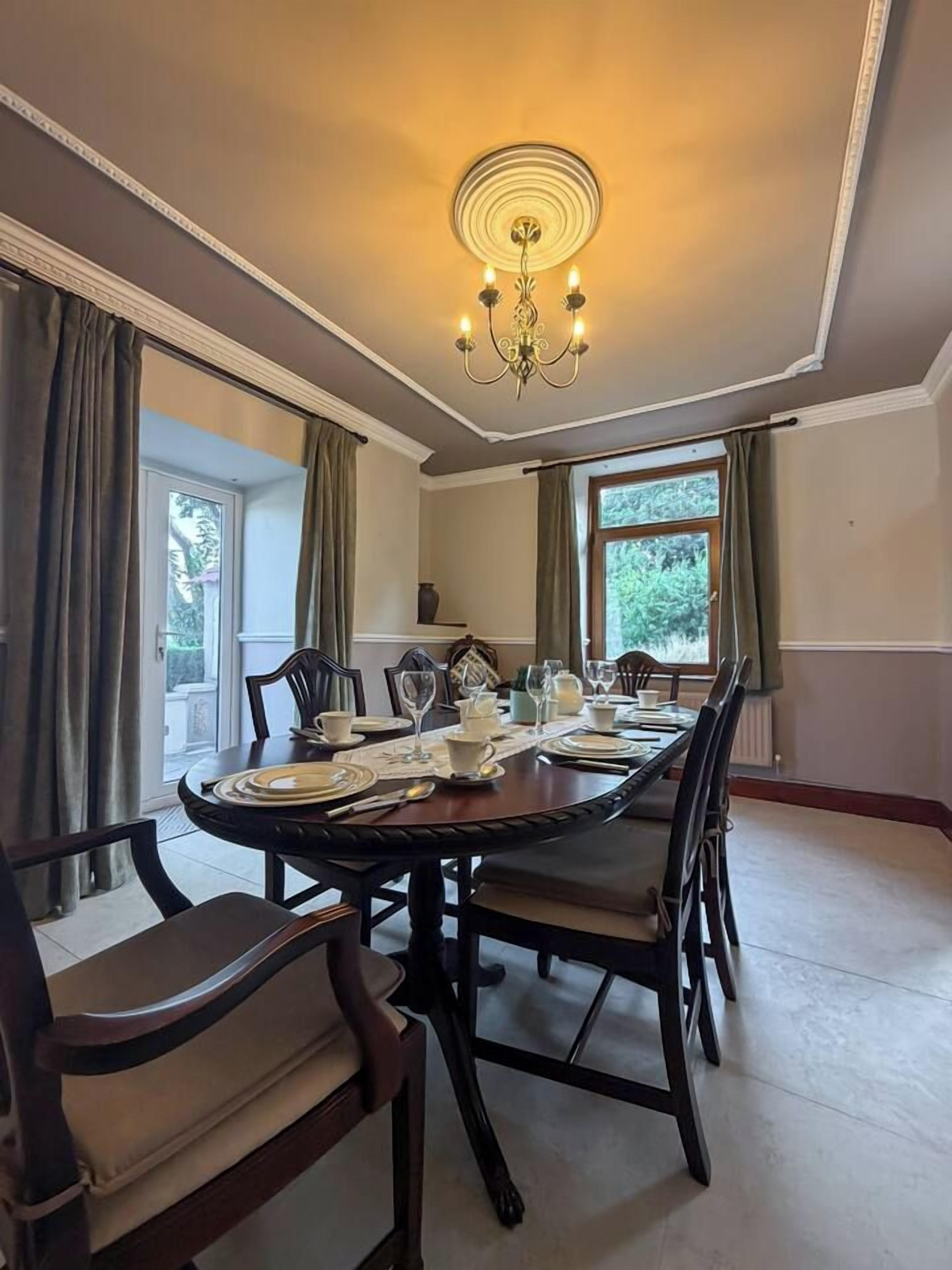 A dining room with a round table set for four, surrounded by chairs, a chandelier, and large windows with curtains.