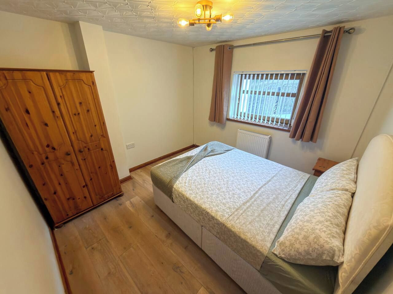 Ein Schlafzimmer mit einem Bett, einem Holzschrank, einem Fenster mit Vorhängen und einer Heizung.