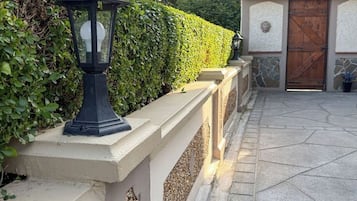 A paved pathway leading to a building with a wooden door, a black lantern on a wall, and a hedge.