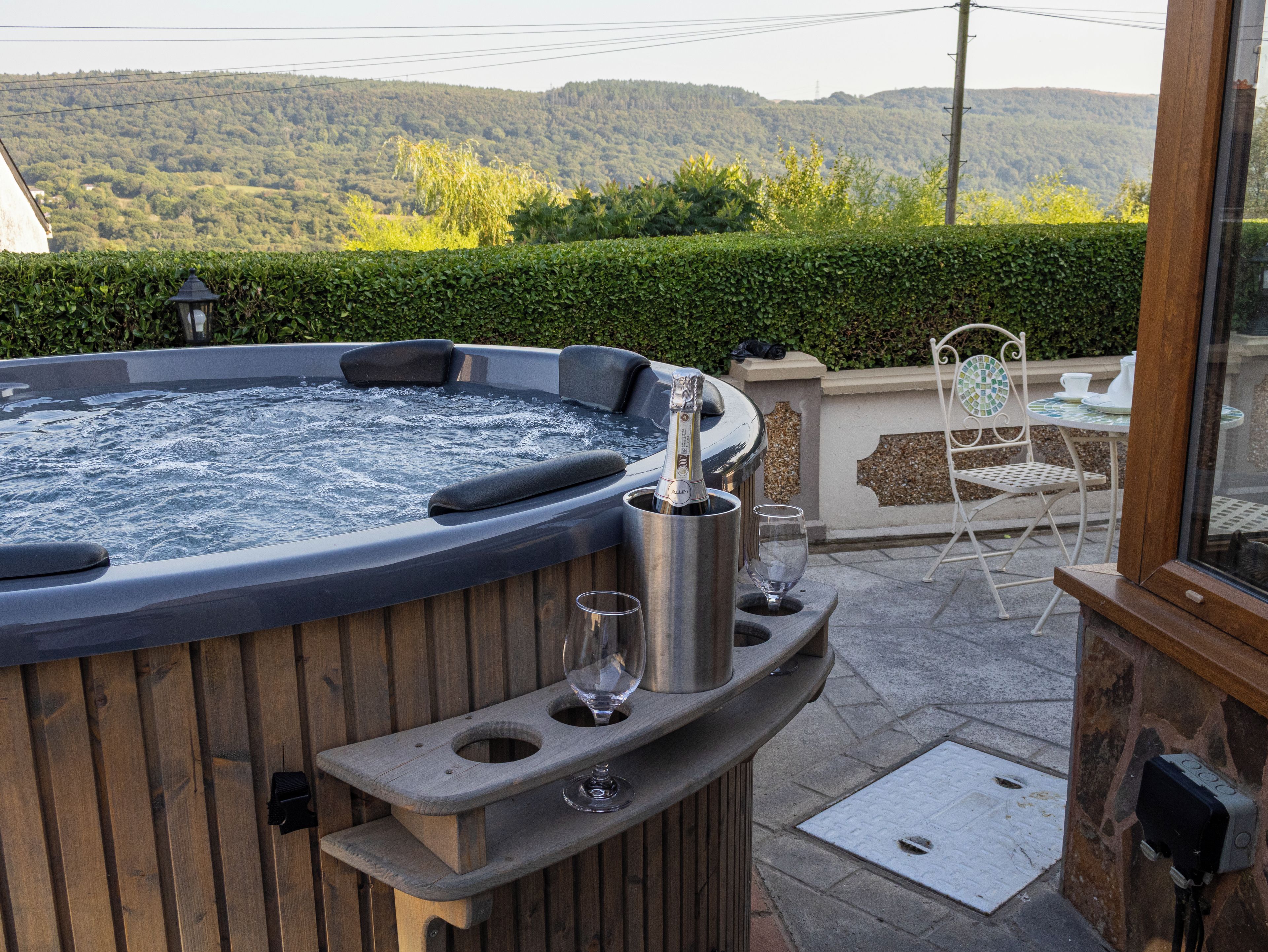 A hot tub with a bottle of champagne, a glass, and a chair on a patio with a view of hills.