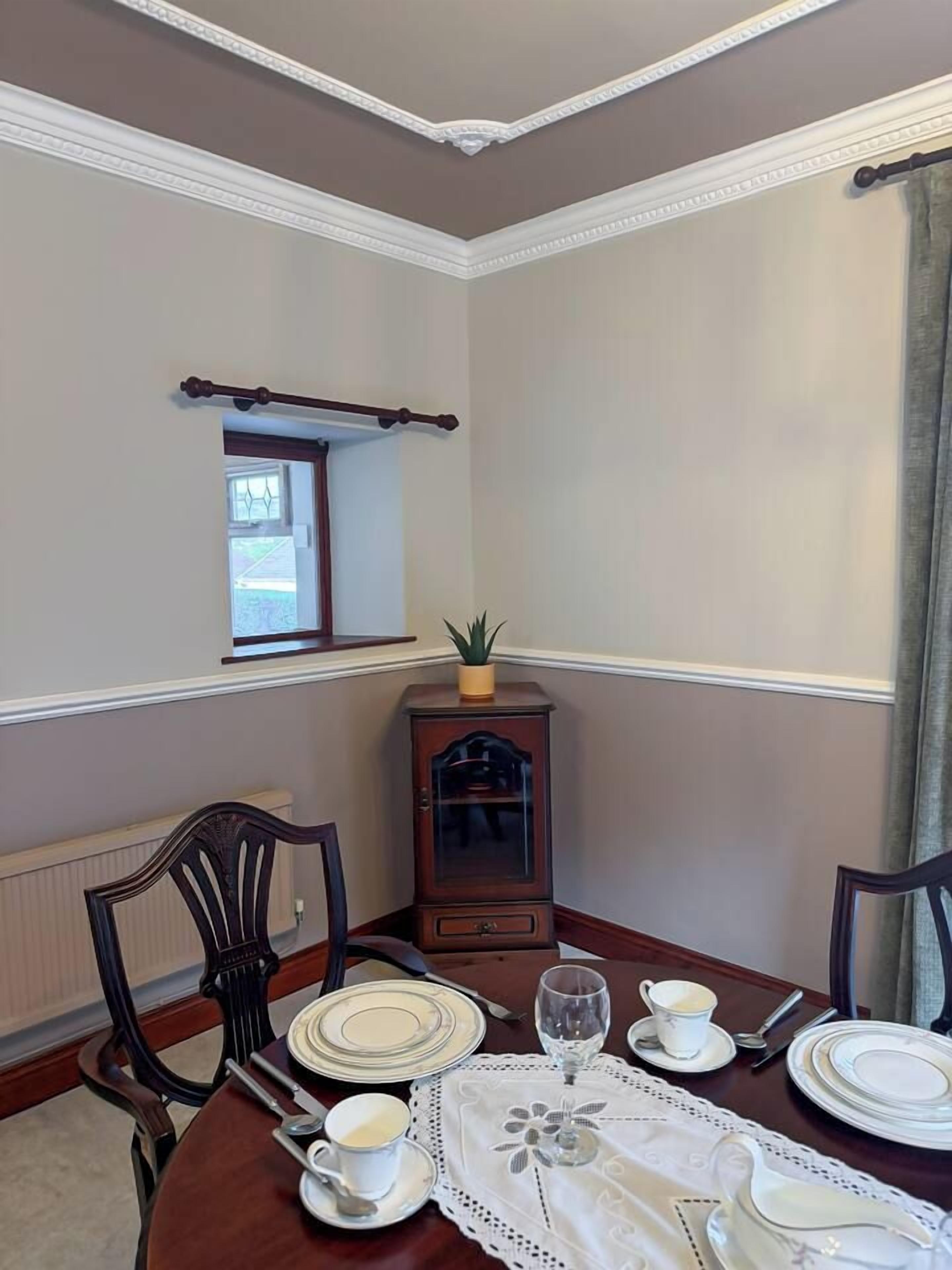 A dining room with a round table set for four, a cabinet with a plant, and a window with curtains.