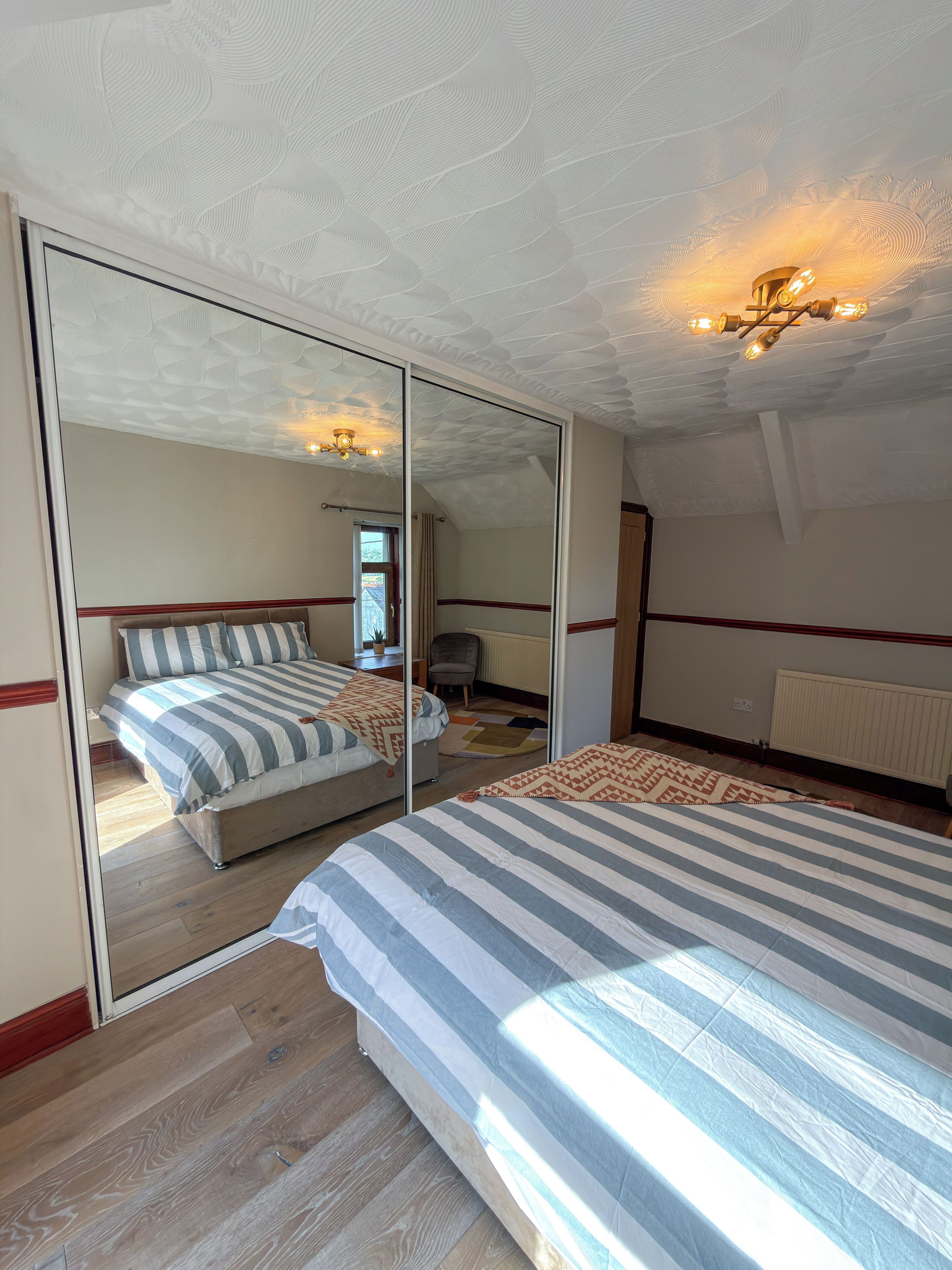 A bedroom with a large mirror, a bed with striped bedding, and a ceiling light fixture.