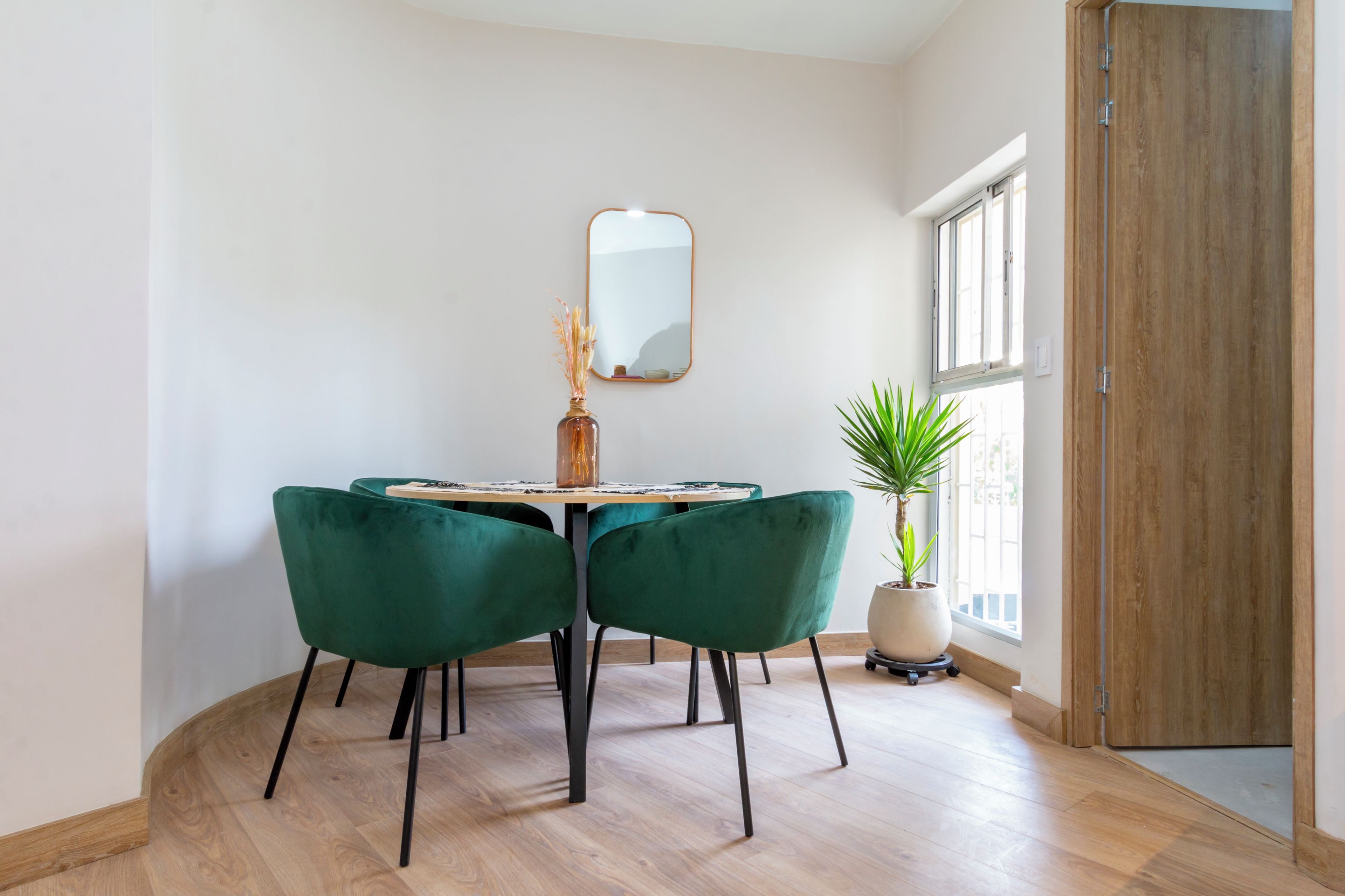 A dining area with a round table, two green chairs, a vase with dried flowers, a mirror on the wall, and a potted plant by the window.