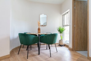 Un comedor con una mesa redonda, dos sillas verdes, un jarrón con flores secas, un espejo en la pared y una planta en maceta junto a la ventana.