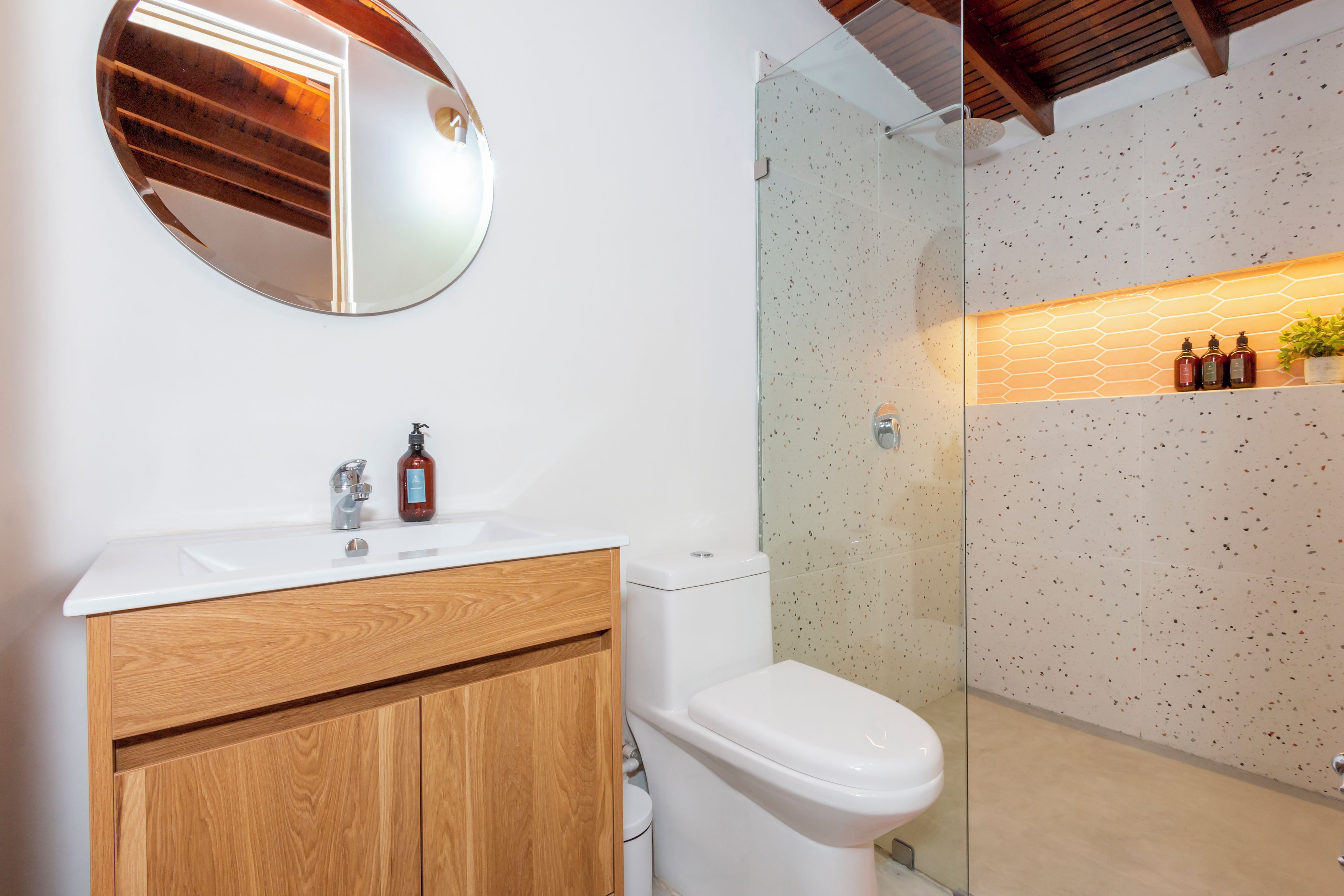 Un baño con tocador de madera, un lavamanos blanco, un espejo redondo, un inodoro y una zona de ducha con pared moteada.