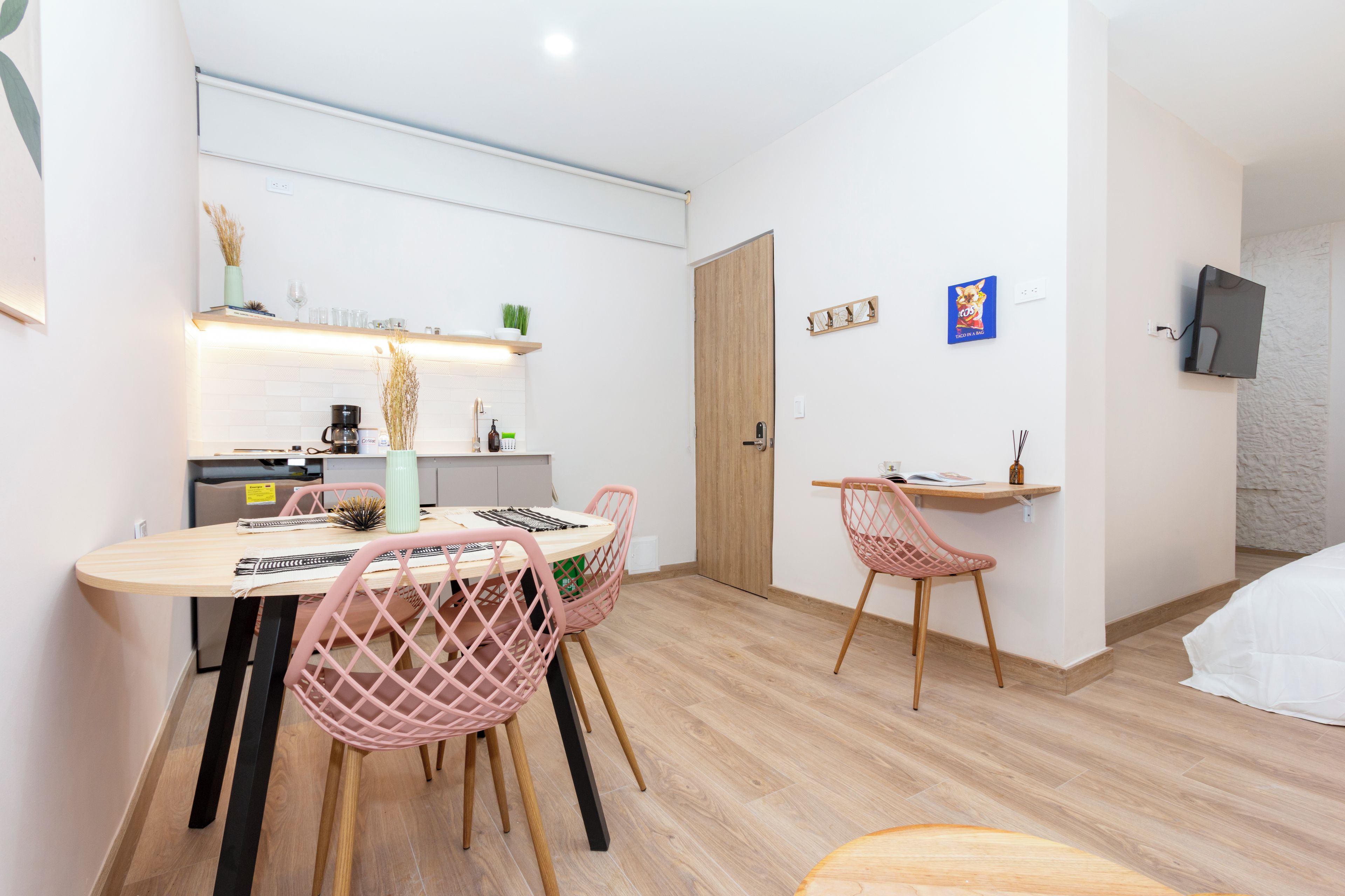 Una habitación moderna y luminosa con una mesa de comedor de madera, sillas rosadas, una pequeña cocina y una cama con ropa blanca.