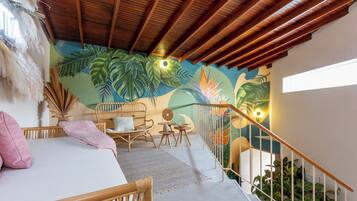 A room with a bed, a wicker chair, a small table, and a staircase with a railing.