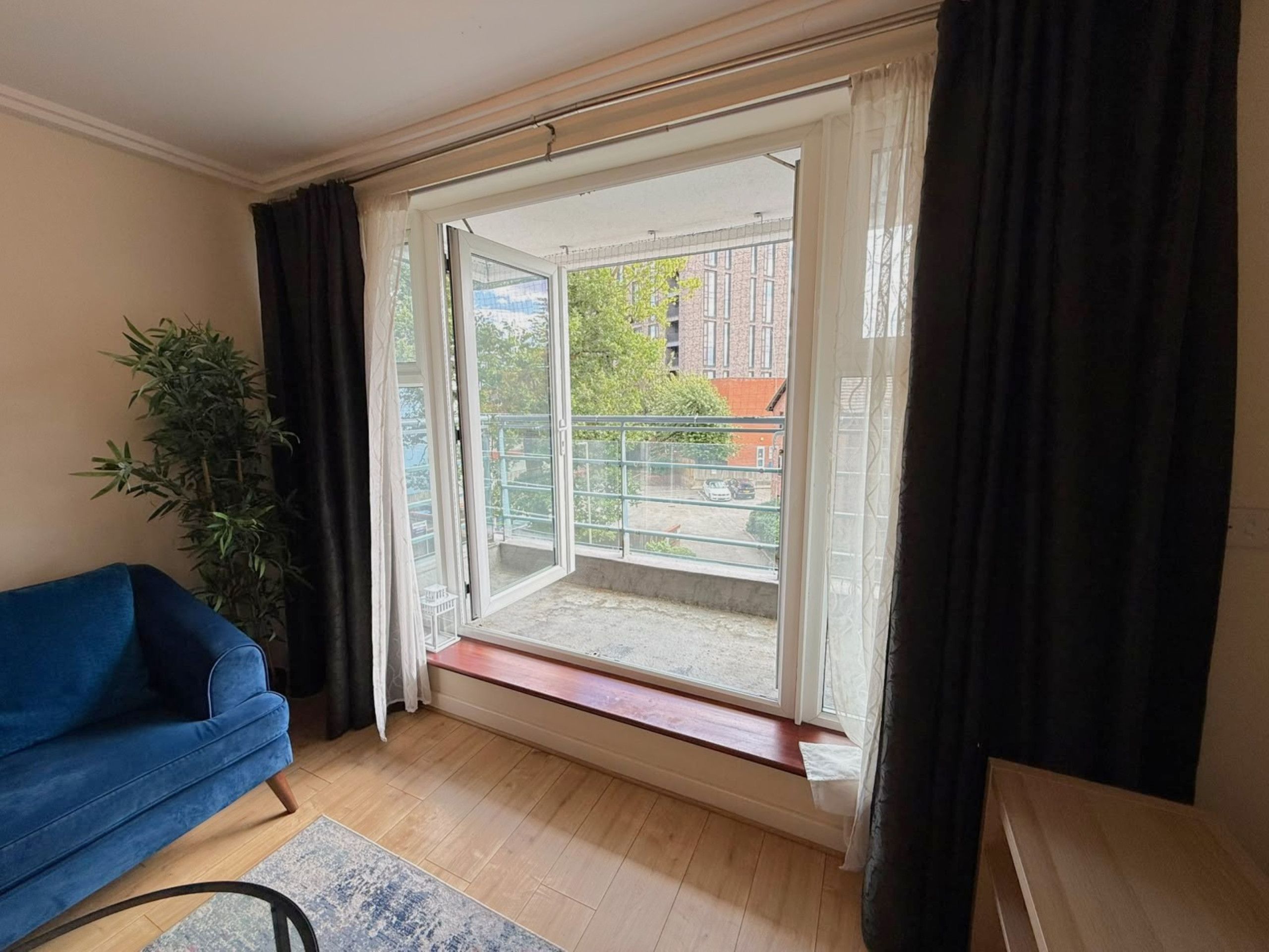 A room with a blue sofa, a potted plant, a sliding glass door leading to an outdoor area with a view of buildings and a fence.