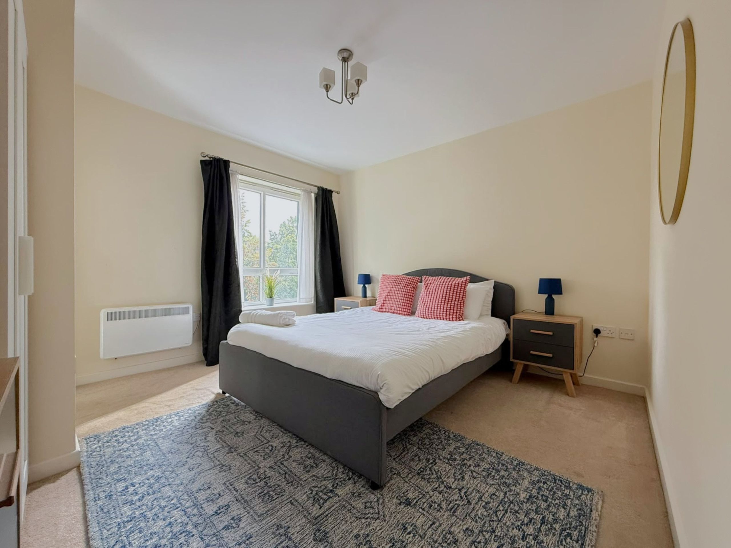 A bedroom with a bed, bedside tables, a window with curtains, a radiator, and a ceiling light fixture.