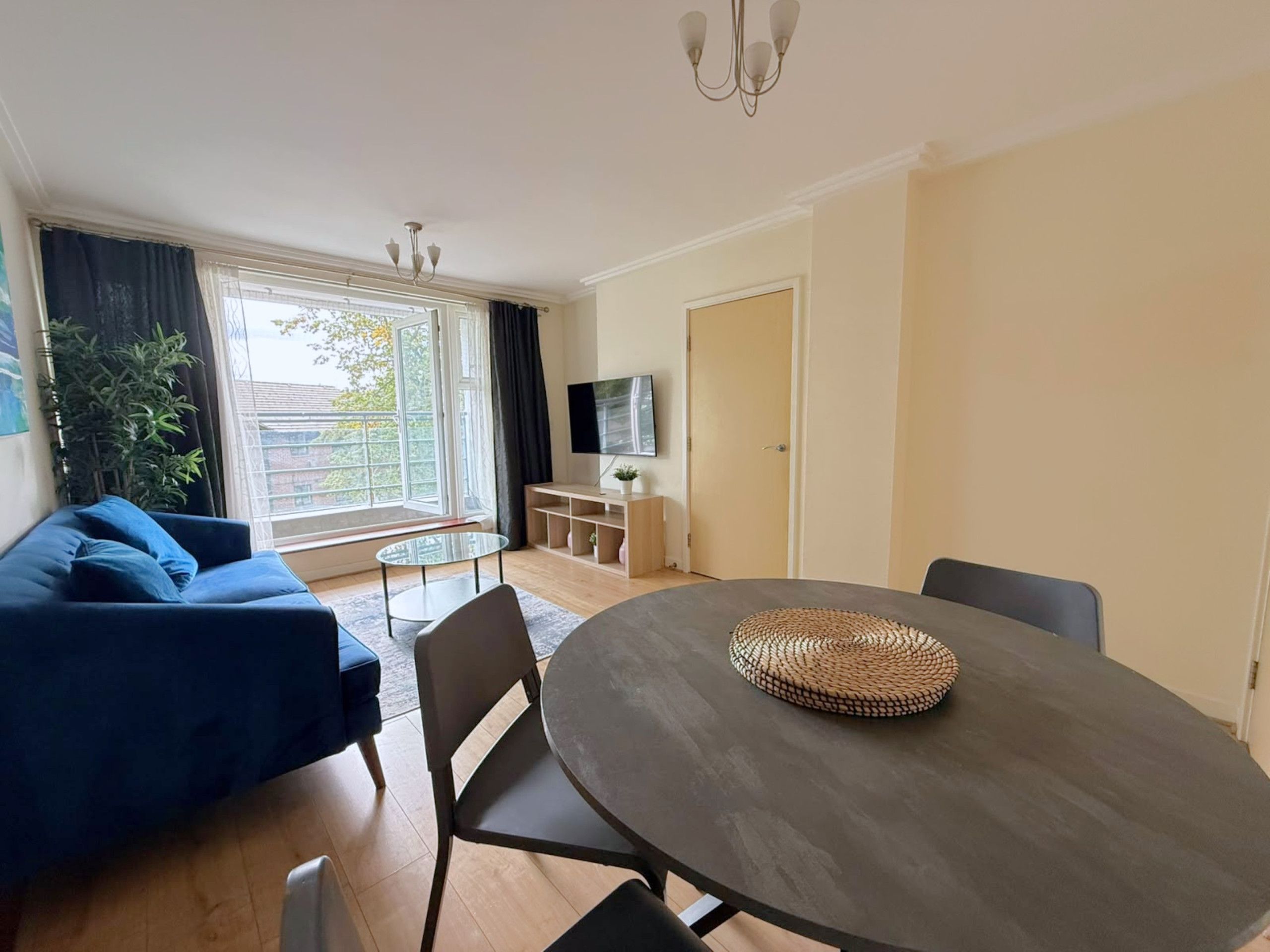 A modern living room with a sofa, a round dining table, chairs, a TV, and a large window with curtains.