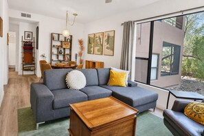A modern living room with a grey sectional sofa, a wooden coffee table, and a view of a building through large windows. - Catalina Cozy, Foothills Retreat with Screened Patio (Tucson)