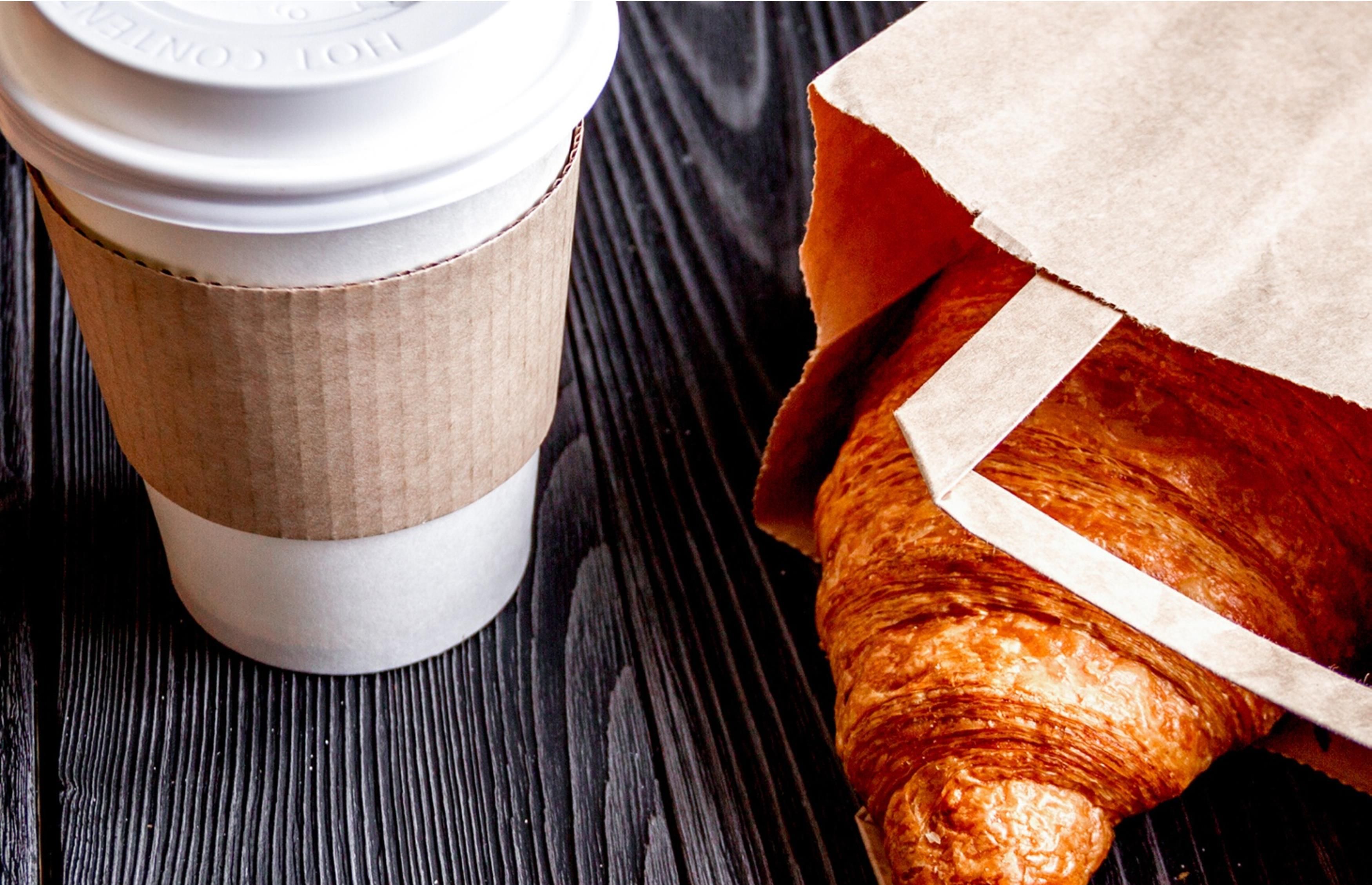 A coffee cup with a lid and a paper bag containing a croissant on a wooden surface.