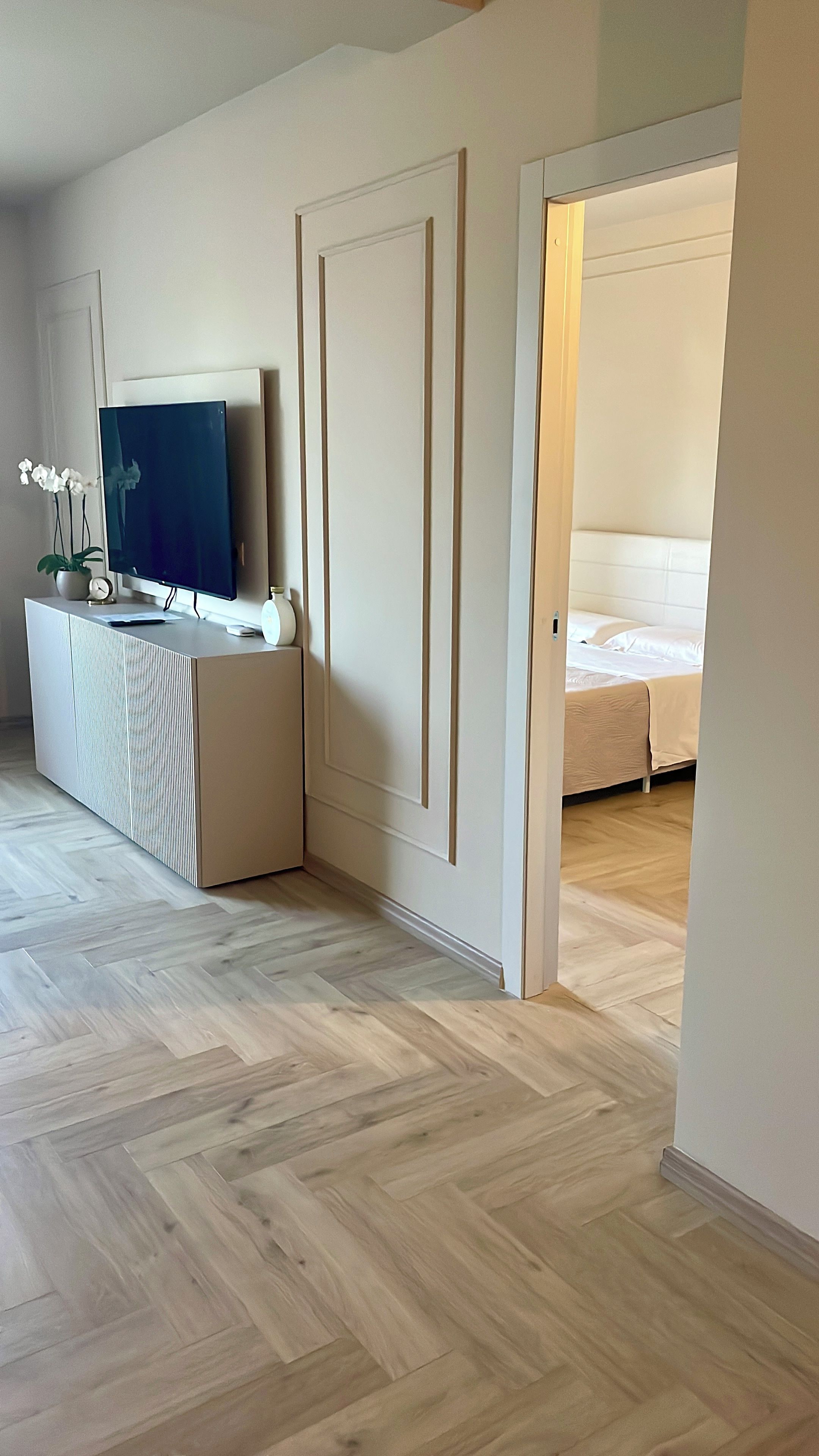 A room with a herringbone-patterned wooden floor, a flat-screen TV on a white cabinet, and a bed with a white headboard.