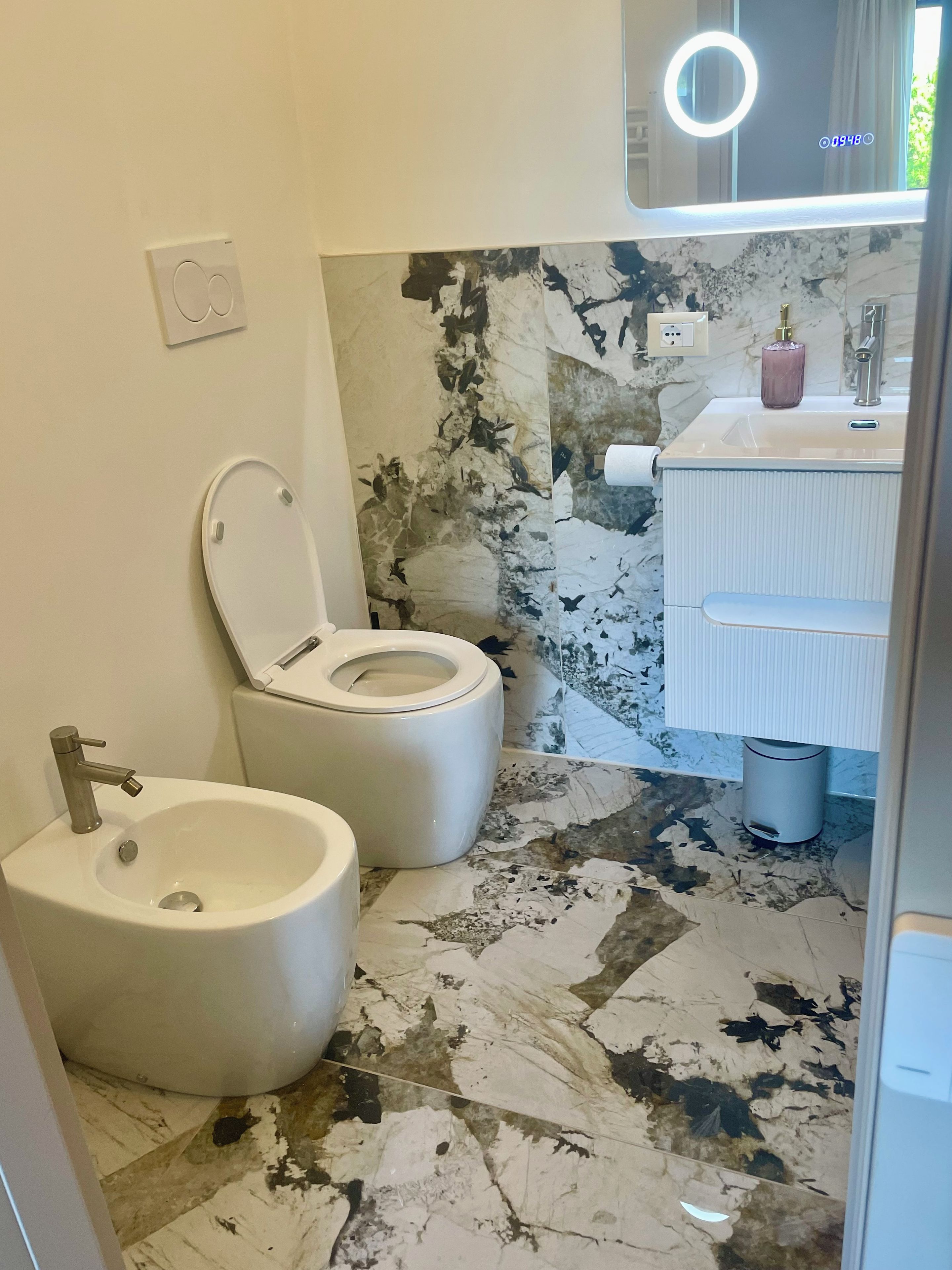 A bathroom with a toilet, bidet, and a sink with a marble backsplash.