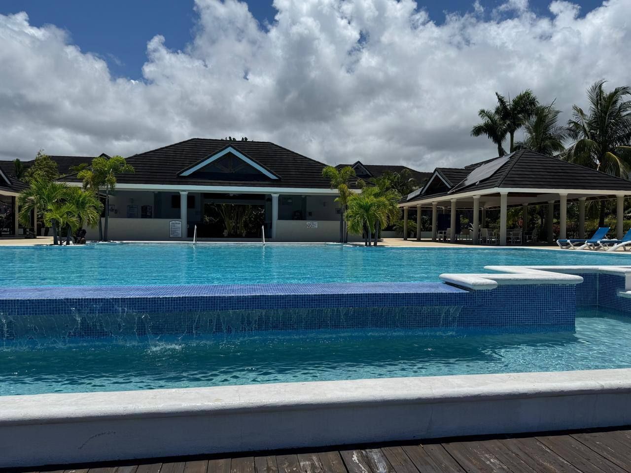 Una piscina con borde de azulejos, rodeada de palmeras y estructuras cubiertas.