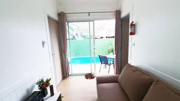 Una habitación con un sofá, una pequeña mesa con un teclado y altavoces, un extintor y vistas a una piscina a través de una puerta de cristal.