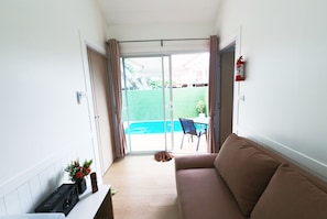 A room with a sofa, a small table with a keyboard and speakers, a fire extinguisher, and a view of a swimming pool through a glass door. - 49 Pool Villa Bangsaen (Mueang Chonburi)