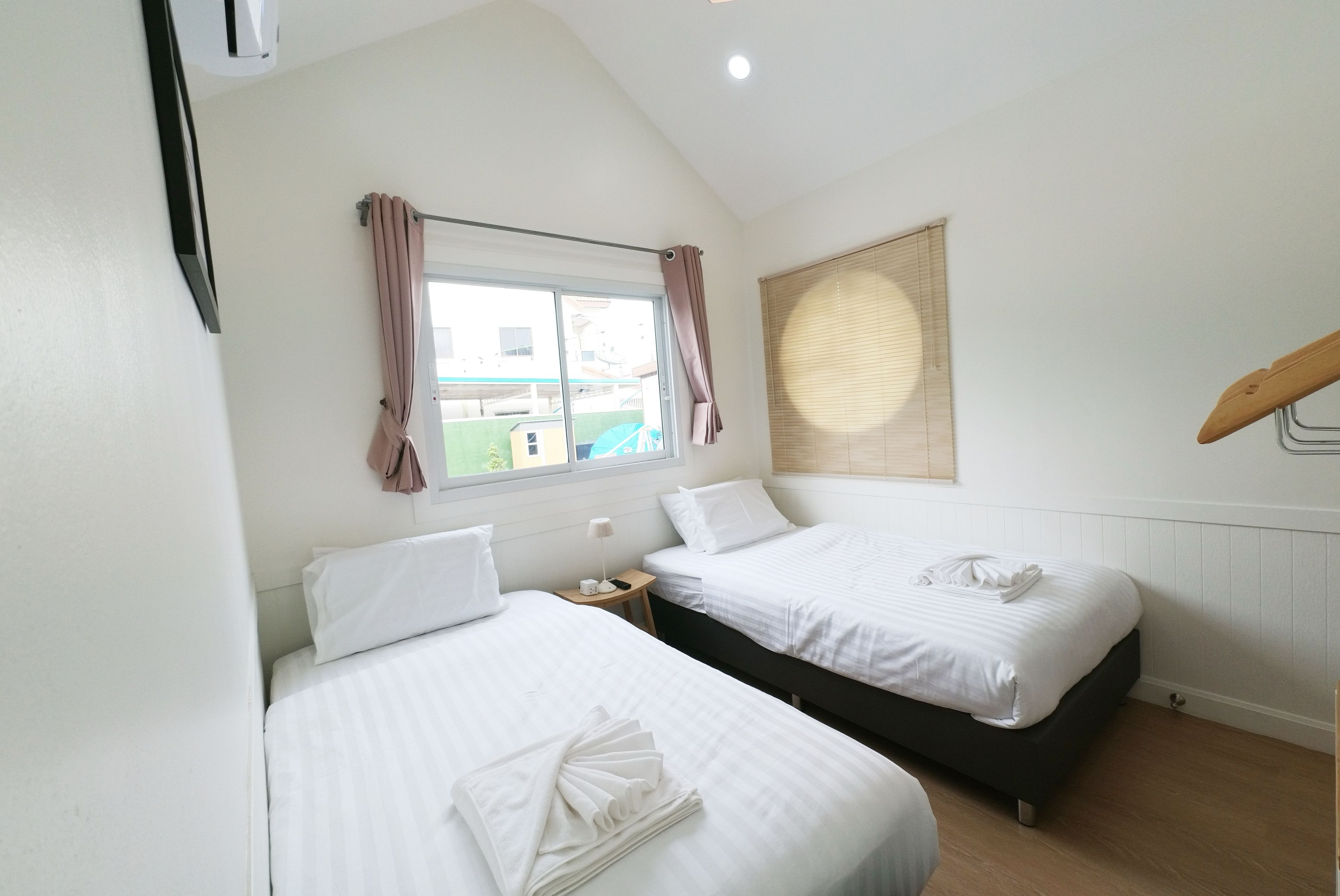 A bedroom with two beds, a window with curtains, a bedside table with a lamp, and a view of the outside.
