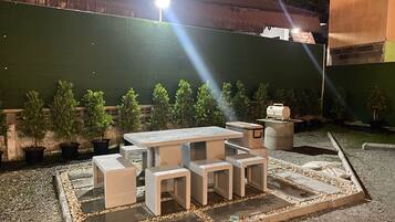 Un área de estar al aire libre con una mesa y bancos, rodeada de plantas en macetas.