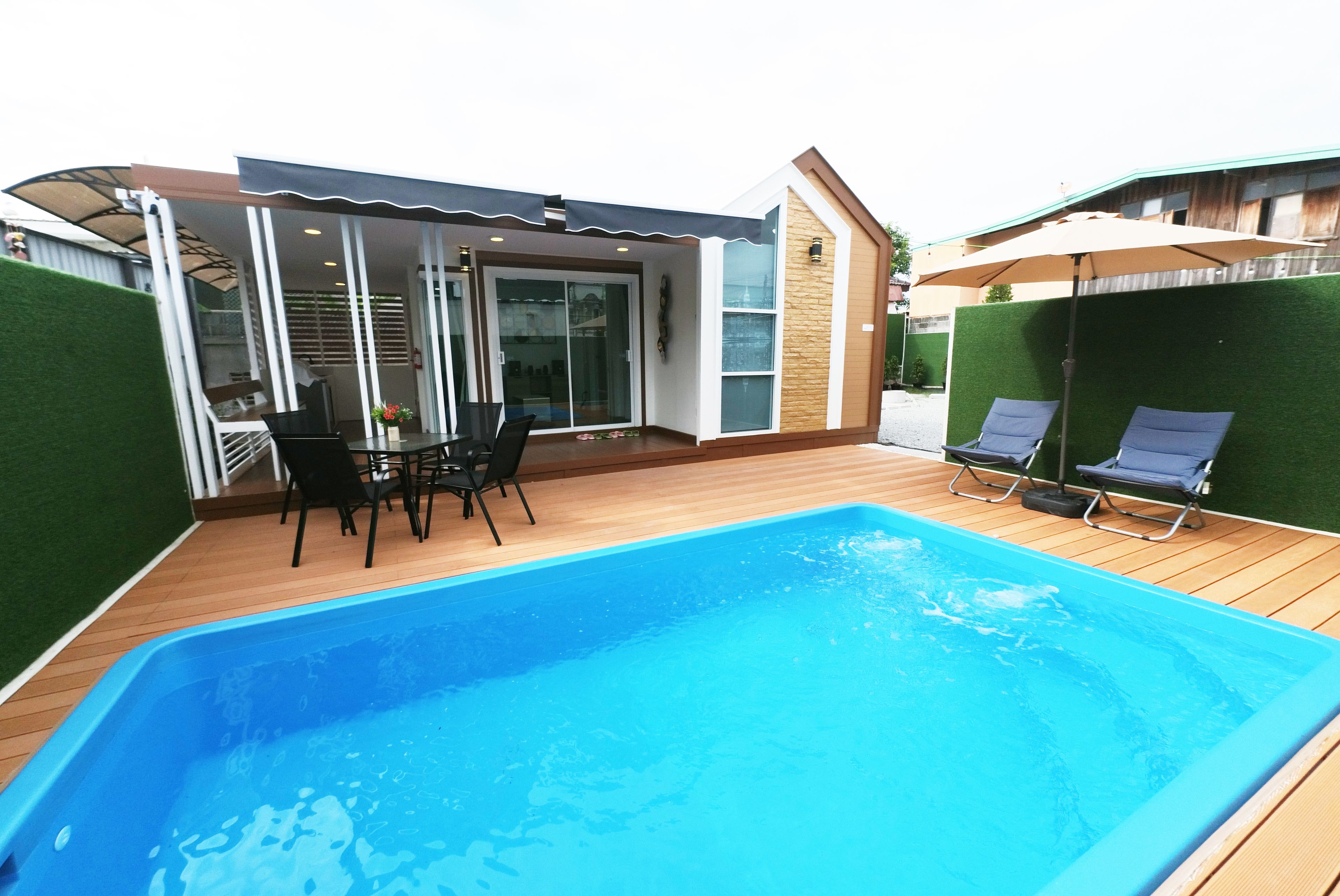 Una piscina con terraza de madera, un área de patio techada y dos sillas de descanso.