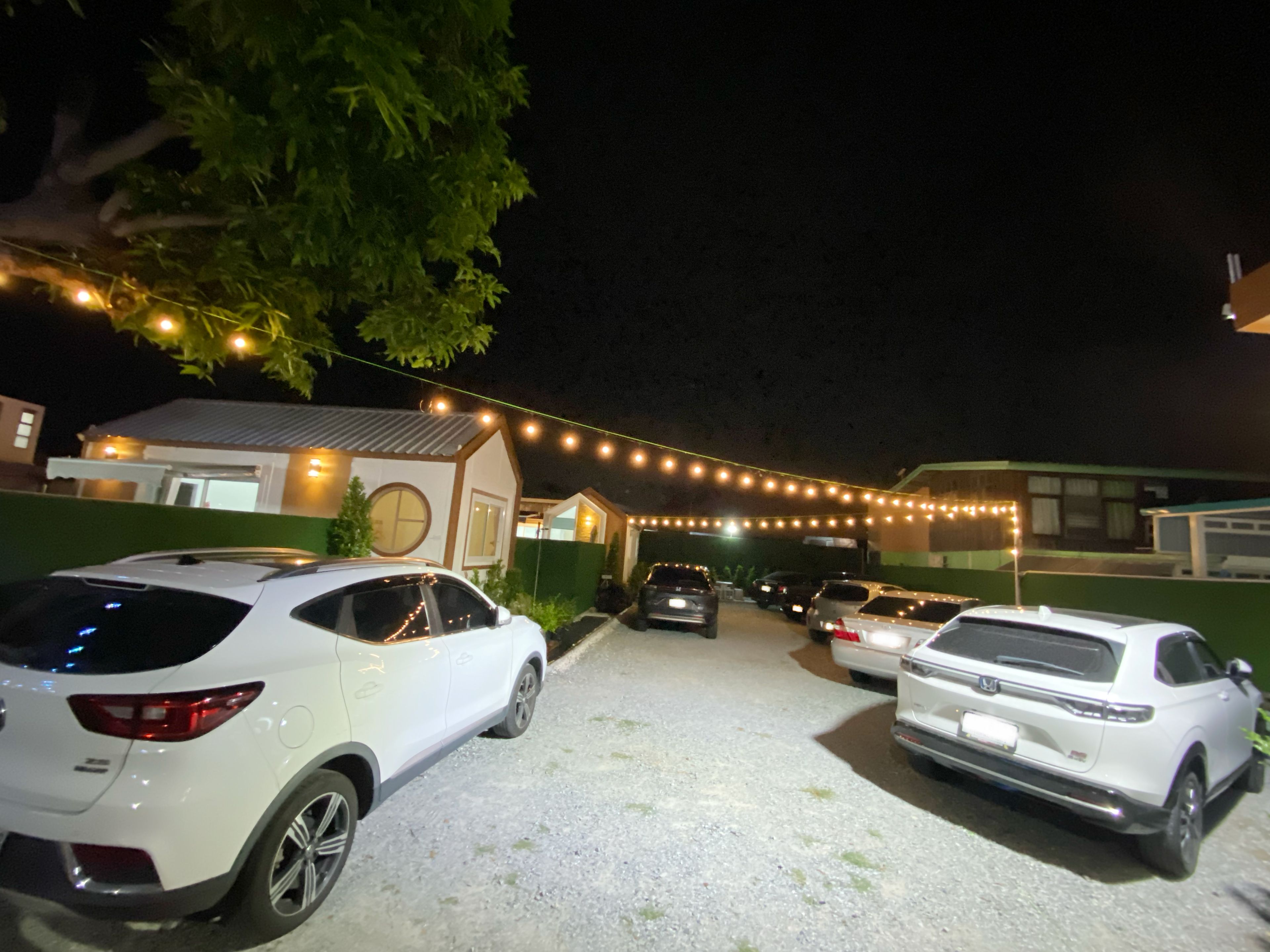 Un estacionamiento con varios autos, un edificio con puerta y ventanas visibles, y luces colgantes.