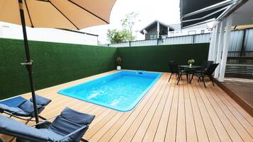 A swimming pool with lounge chairs and a table, surrounded by a wooden deck and a green wall.