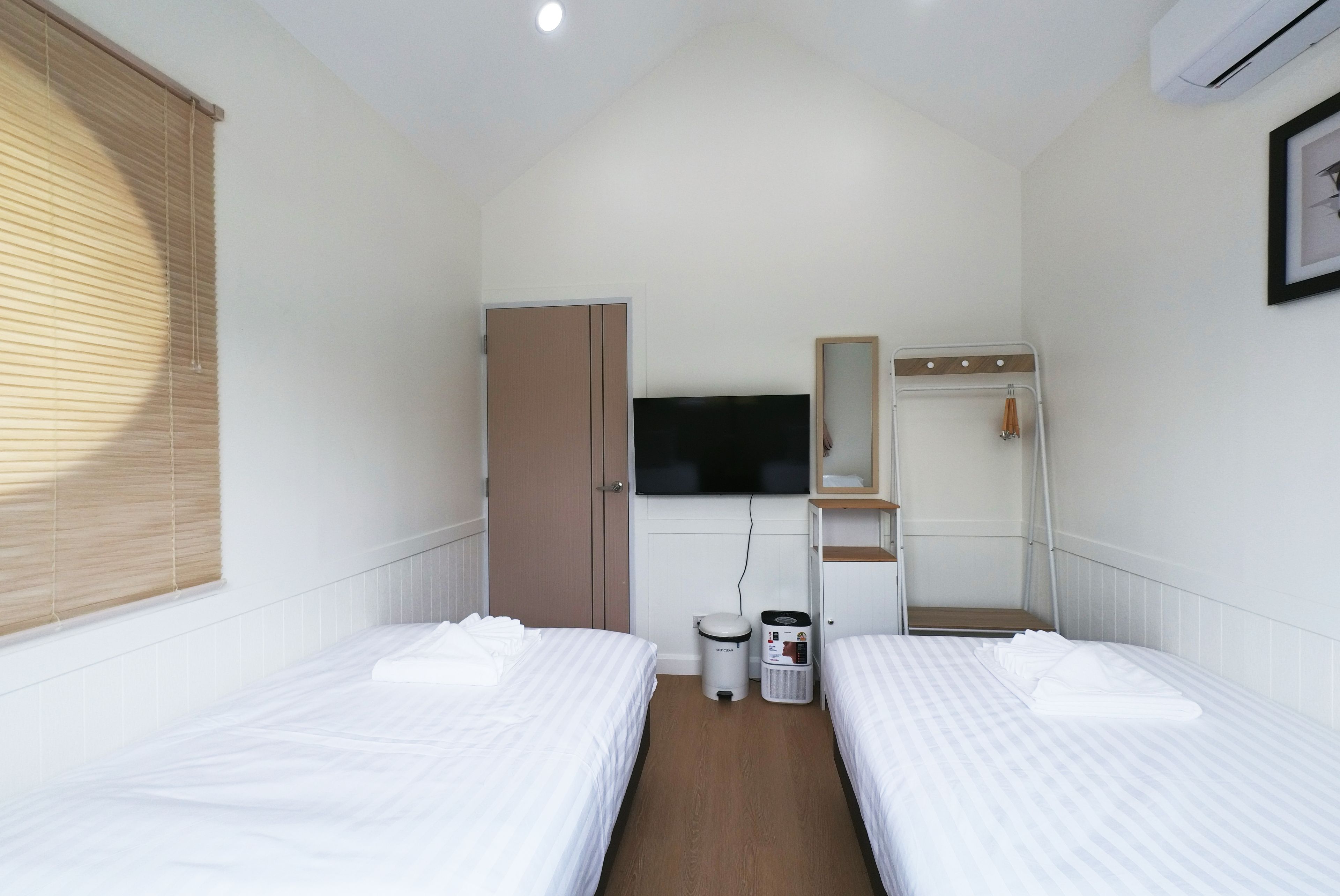 A hotel room with two single beds, a wardrobe, a television, a mirror, a waste bin, and an air conditioning unit.