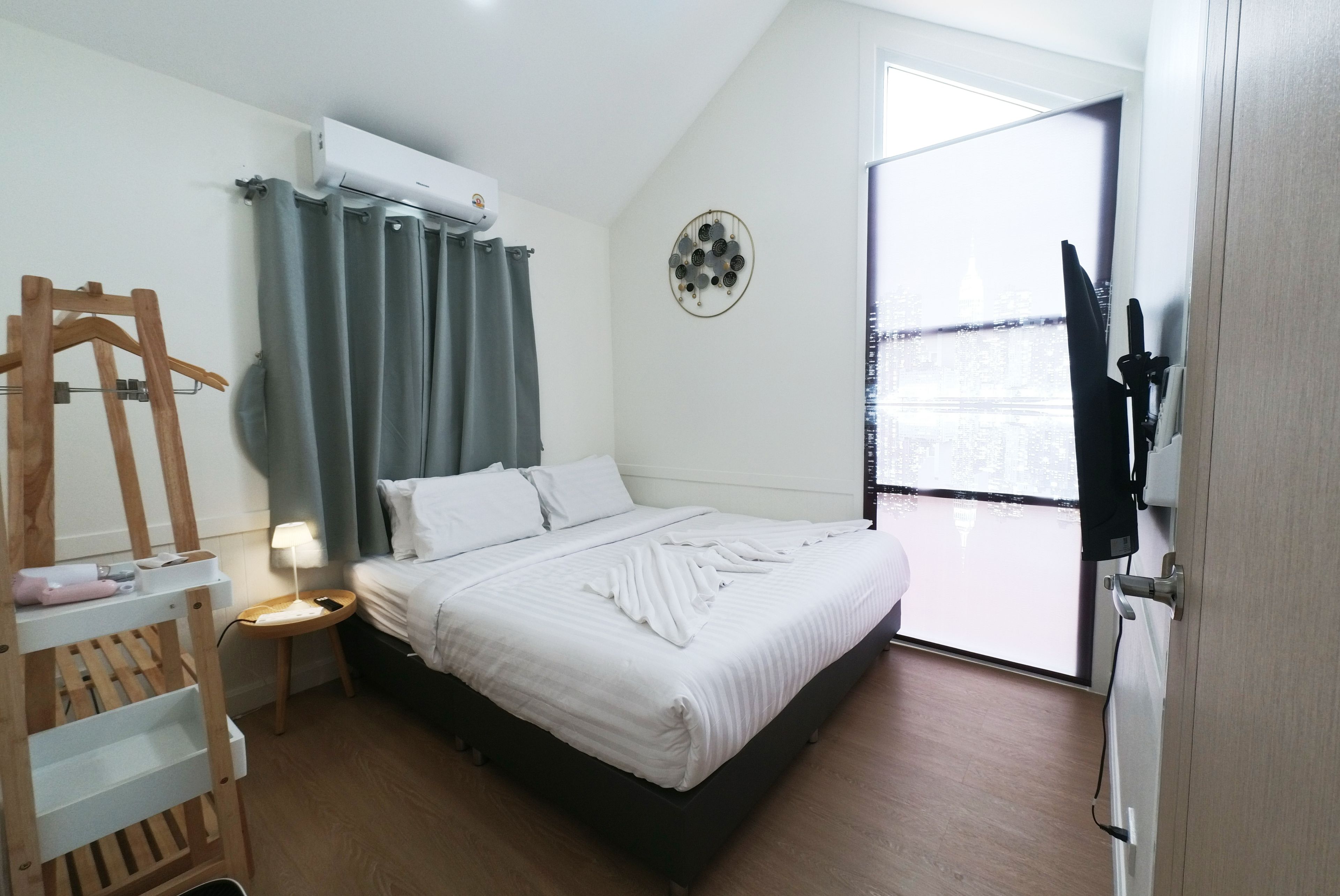 A hotel room with a bed, a bedside table, a hanging coat rack, a wall-mounted air conditioner, and a view of the city through the window.