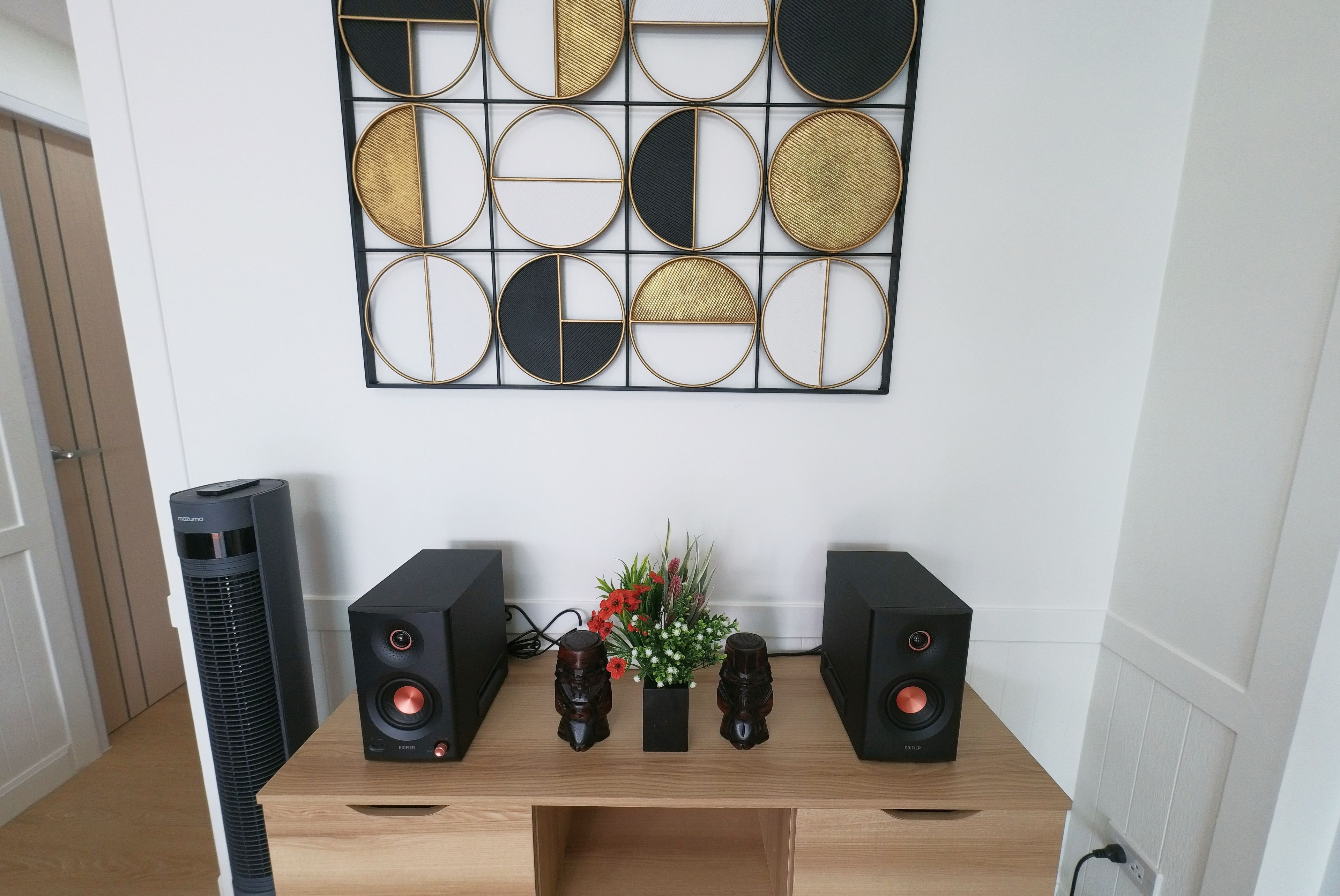 A room with a wooden console table, two black speakers, a decorative wall piece, and a tall black air purifier.