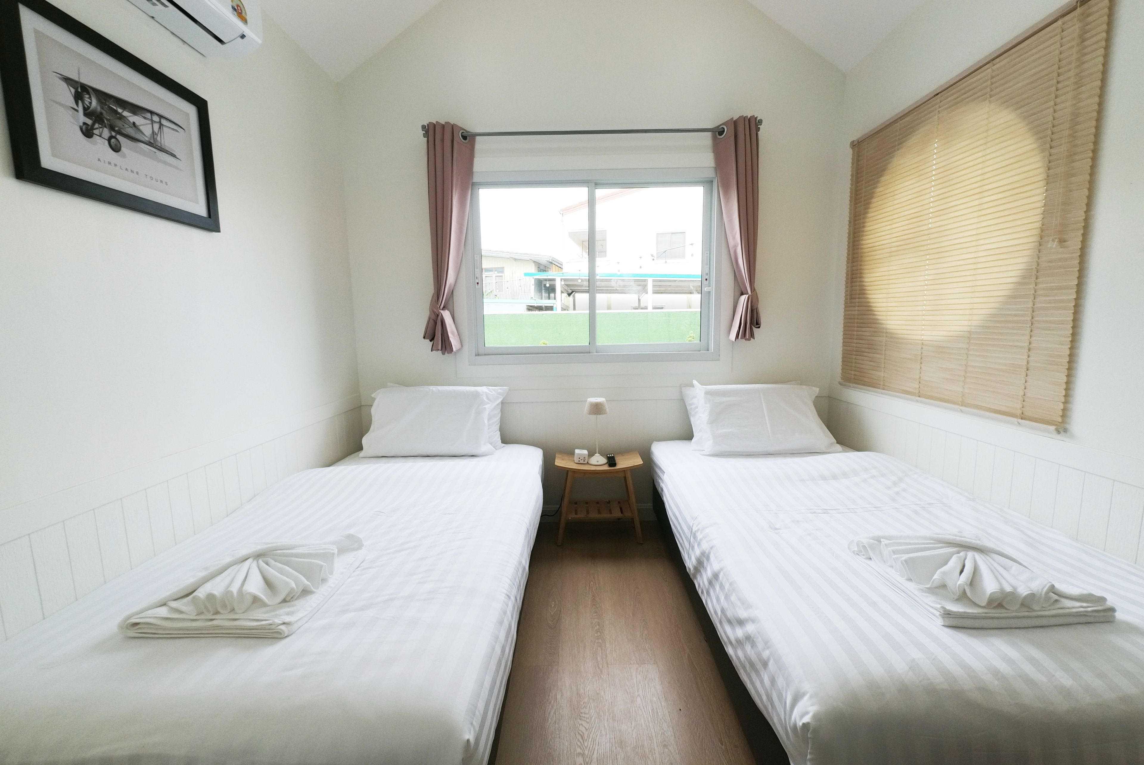 Habitación de hotel con dos camas individuales, una ventana con cortinas, una mesita de noche pequeña con lámpara y un cuadro en la pared.