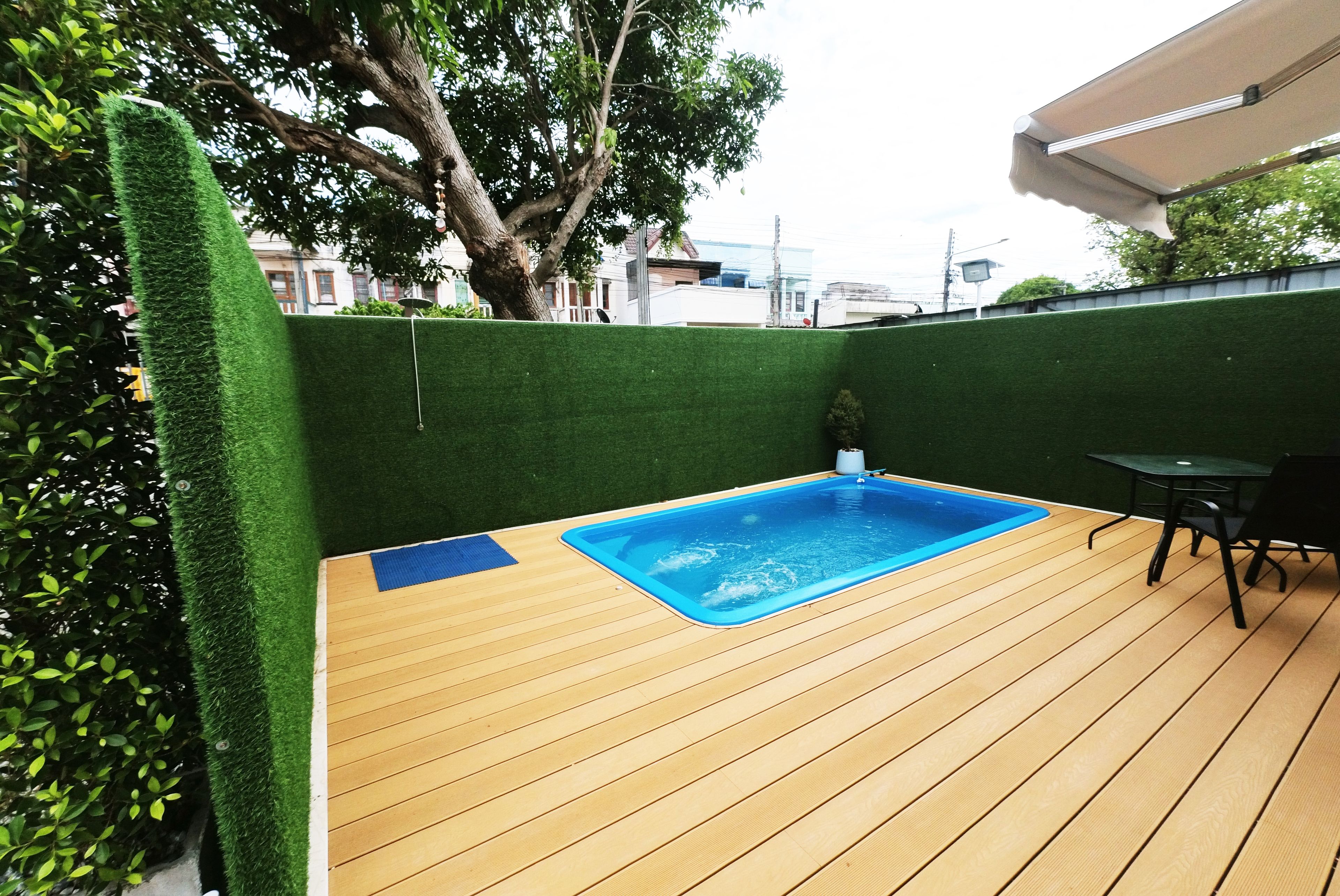 Una terraza de madera con piscina, rodeada de césped artificial verde y un árbol.