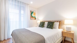 Ein Hotelzimmer mit einem großen Bett, einem Nachttisch mit Lampe und einem Fenster mit durchsichtigen Vorhängen.