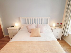A bedroom with a bed, two bedside tables, a nightstand, a window with curtains, and a wall-mounted lamp. - Apartment 17, St. John's (Whitby)