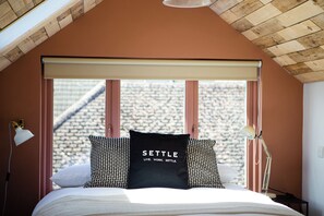 Ein Schlafzimmer mit geneigter Decke, einem Bett mit weißer Bettwäsche, zwei Nachttischlampen und einem Fenster mit Blick auf eine Steinmauer.