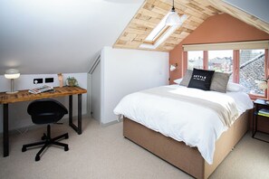 A bedroom with a sloped ceiling, a bed with white bedding, a desk with a chair, and a window with a view of buildings. - Chalet style 1-bedroom studio in charming Southampton perfect for your getaway (Southampton)