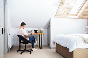 Eine Person sitzt an einem Schreibtisch in einem Zimmer mit einem Bett, einem Heizkörper und einem Oberlicht.