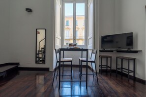 A room with a dining table, two chairs, a TV on a stand, and a window with a view of buildings. - Living Filomarino Collection (Naples)