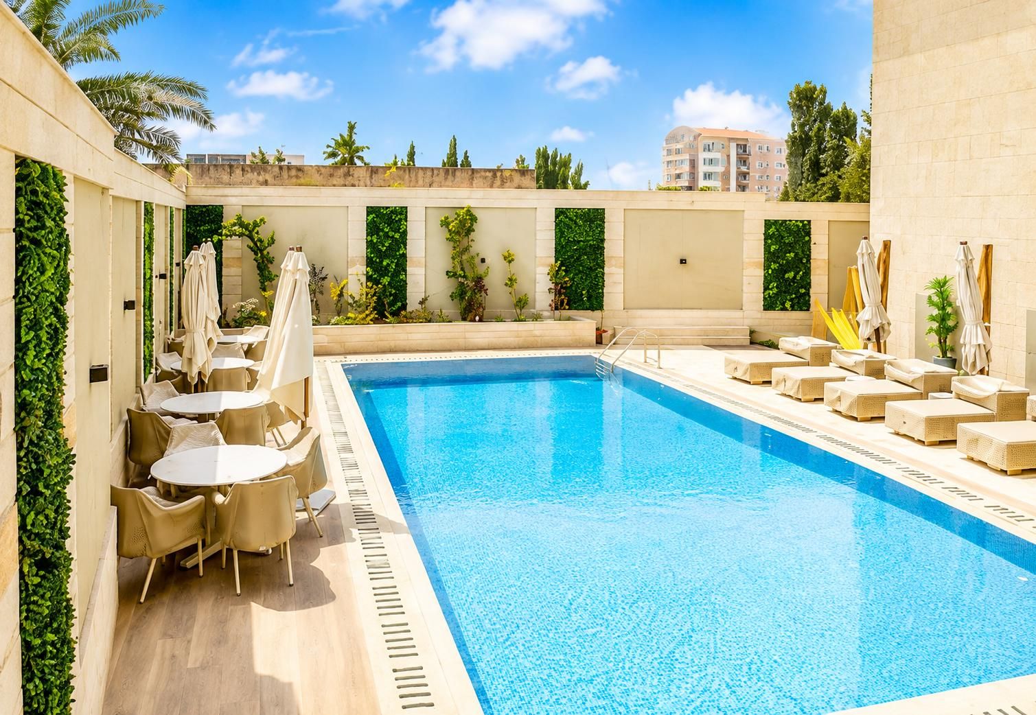 A swimming pool with lounge chairs and tables, surrounded by greenery and a clear sky.