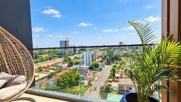 Ein Balkon mit Korbstuhl und Blumentopf, mit Blick auf eine Stadtlandschaft mit Gebäuden und Grünflächen.