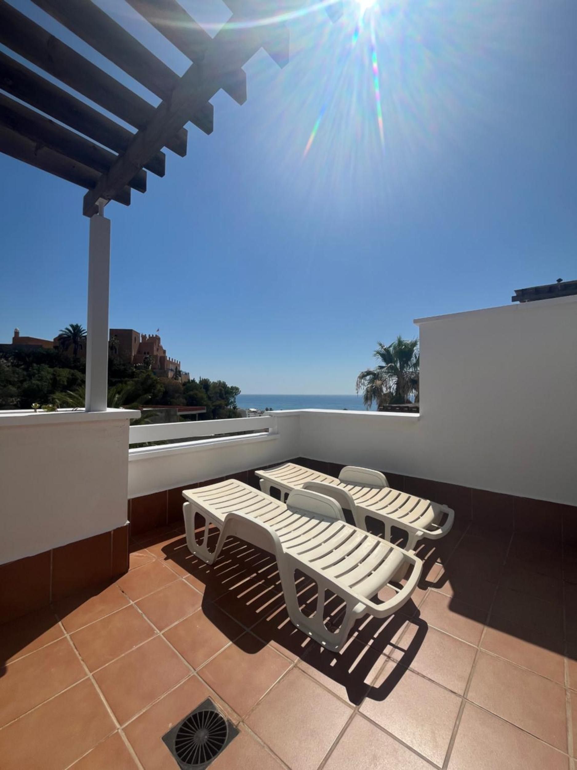 Una terraza con vista al mar, que incluye una pérgola, sillas de plástico blanco y una mesa.