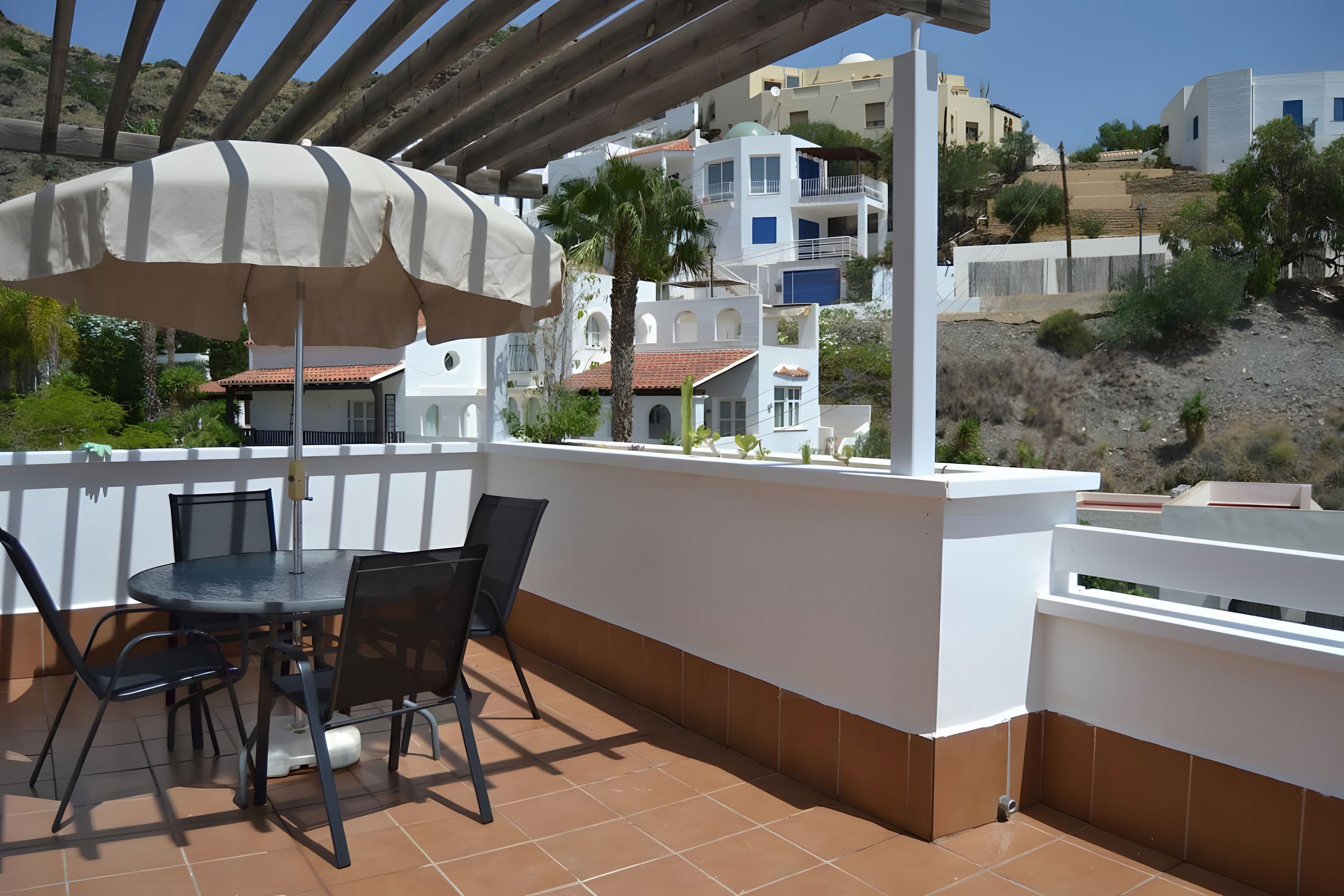 An outdoor patio with a table and chairs, a pergola with a canopy, and a view of buildings in the background.