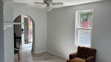 Un salón amplio con ventilador de techo, un sillón marrón, un reposapiés blanco y un ventanal con vistas a una valla.