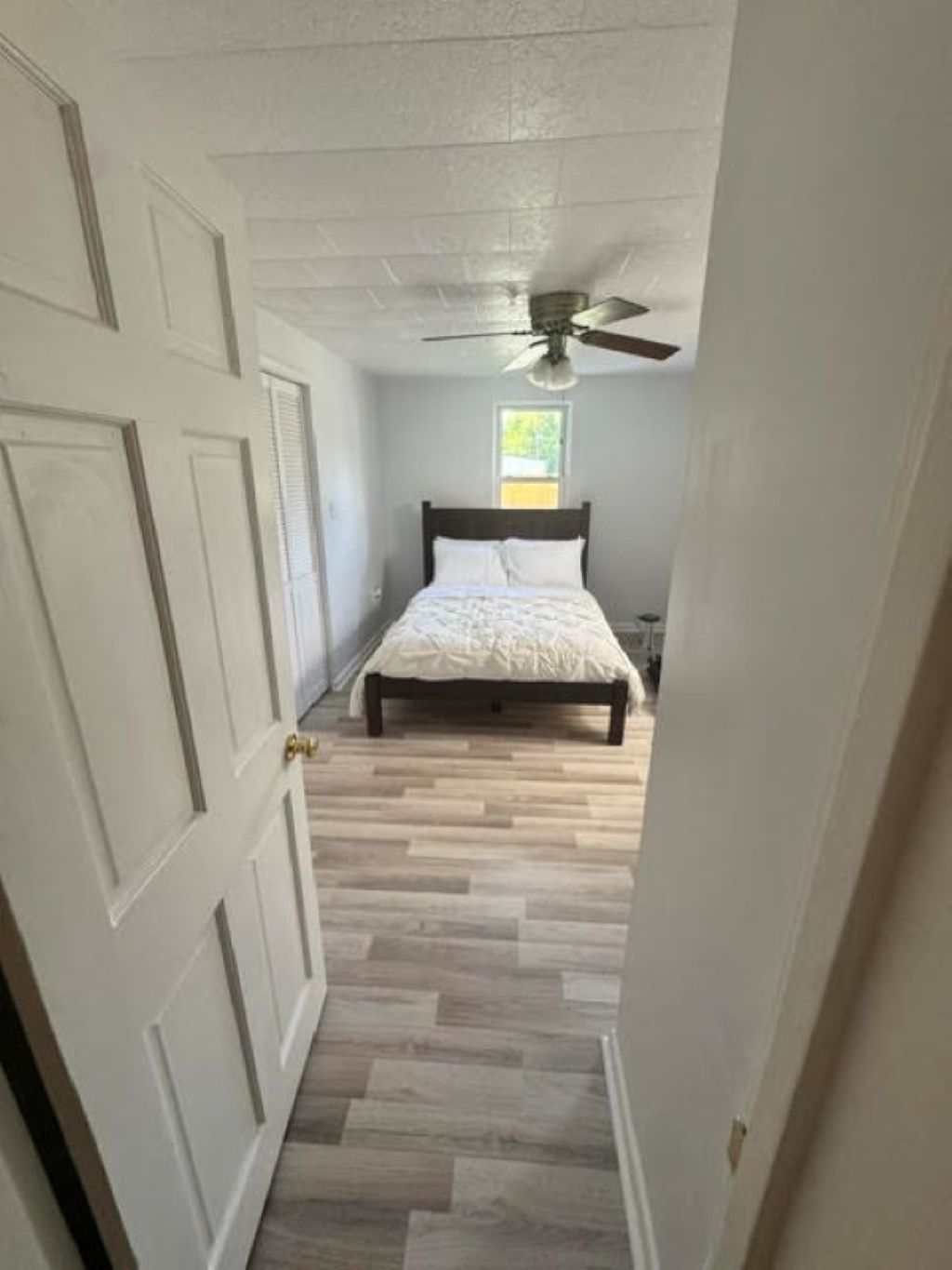 Un couloir étroit mène à une chambre à coucher qui contient un lit, un ventilateur de plafond et une fenêtre.